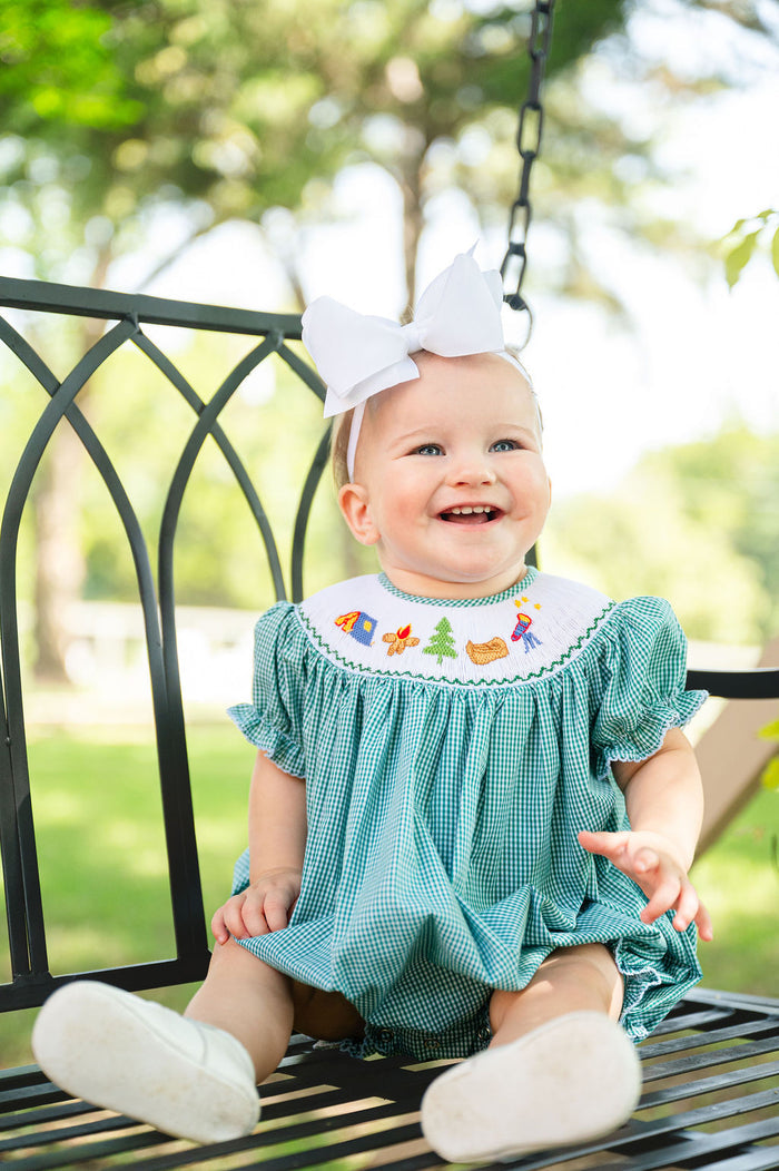 Baby girl sitting on porch swing wearing Smocked Camping Bubble by Ruth and Ralph, white bow, outdoors