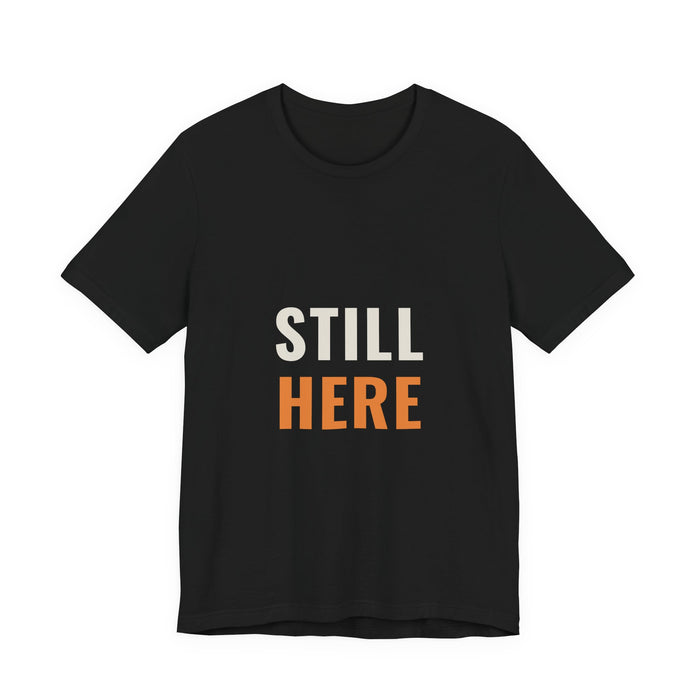 STILL in cream capitals above HERE in orange, printed on a black unisex t-shirt.
