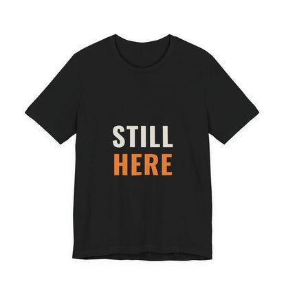 STILL in cream capitals above HERE in orange, printed on a black unisex t-shirt.