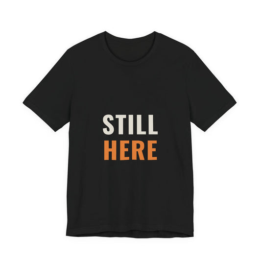 STILL in cream capitals above HERE in orange, printed on a black unisex t-shirt.